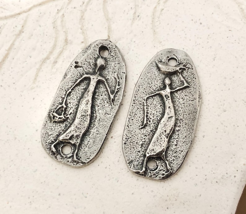 Peut inclure: Deux pendentifs argent&eacute;s de forme ovale, perc&eacute;s d'un trou en haut et en bas. Chaque pendentif repr&eacute;sente une figure stylis&eacute;e aux bras tendus. L'une tient un objet, l'autre porte une forme de croissant sur la t&ecirc;te.