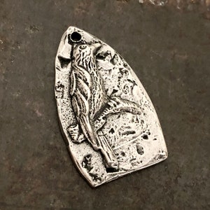 May include: A silver-toned metal pendant shaped like a shield with a pointed top and rounded base. The surface features a raised, textured relief of a bird perched on a branch, with a small circular hole at the top for attachment to a chain or cord.