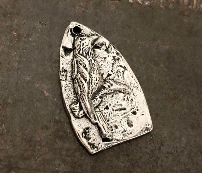 May include: A silver-toned metal pendant shaped like a shield with a pointed top and rounded base. The surface features a raised, textured relief of a bird perched on a branch, with a small circular hole at the top for attachment to a chain or cord.