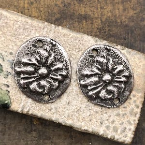 May include: Two antique silver-toned metal oval pendants with a floral design. Each pendant has a textured surface and two holes for stringing. The pendants are displayed on a textured, light-colored surface.