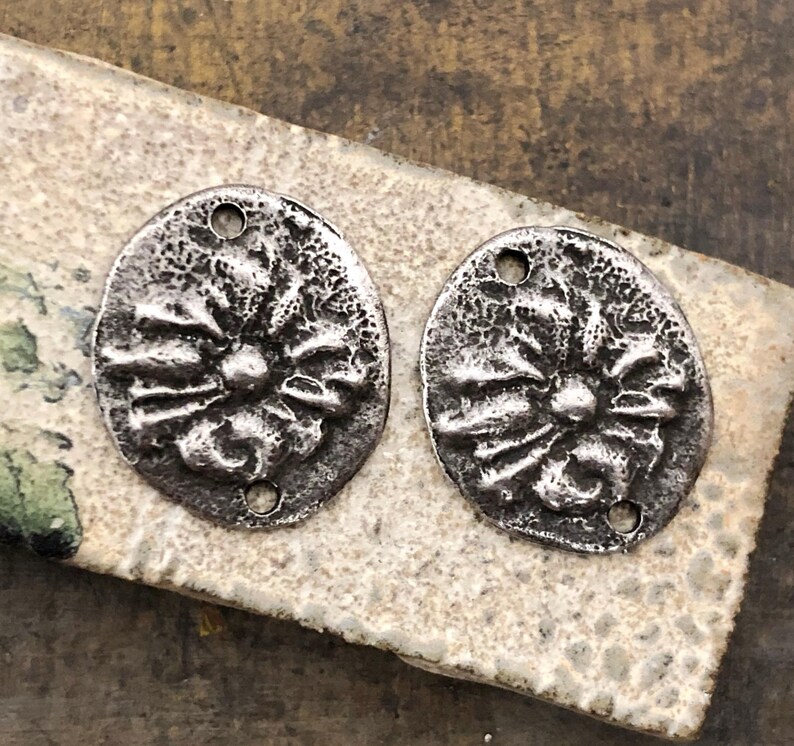 May include: Two antique silver-toned metal oval pendants with a floral design. Each pendant has a textured surface and two holes for stringing. The pendants are displayed on a textured, light-colored surface.