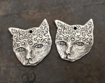 May include: Two silver cat face charms with a swirl pattern. The charms have a hole at the top for attaching to jewelry.
