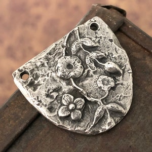 Puede incluir: Un colgante de metal plateado con un dise&ntilde;o floral. El colgante presenta flores, hojas y tallos en relieve. Tiene una superficie texturizada y dos agujeros para colgar. La forma general es un rect&aacute;ngulo redondeado.