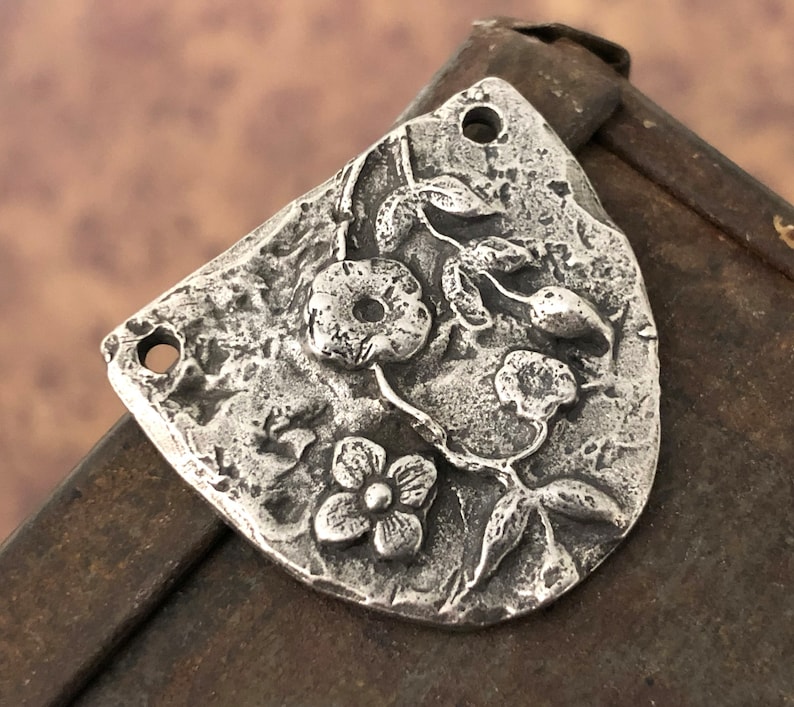 Puede incluir: Un colgante de metal plateado con un dise&ntilde;o floral. El colgante presenta flores, hojas y tallos en relieve. Tiene una superficie texturizada y dos agujeros para colgar. La forma general es un rect&aacute;ngulo redondeado.