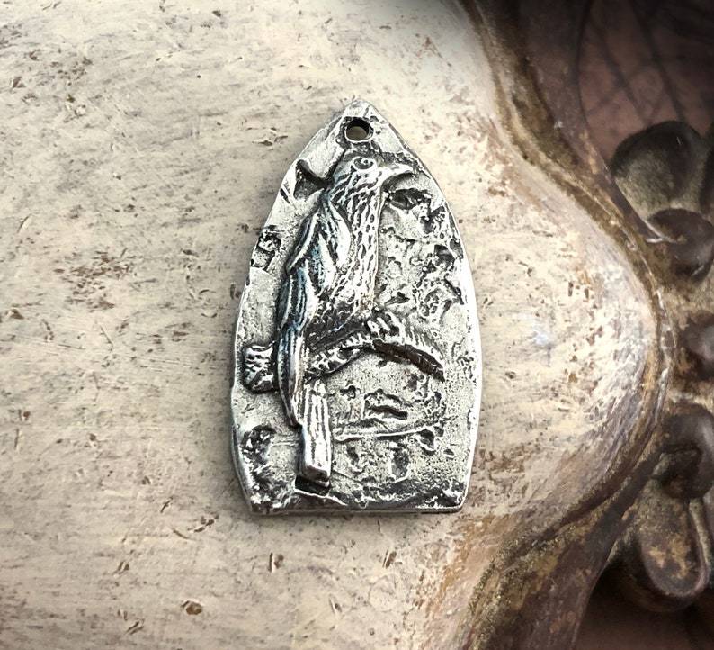 May include: A silver charm in the shape of a bird with detailed etching. The charm is shaped like a teardrop and has a loop at the top for attaching to a necklace or bracelet.