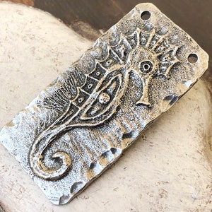 Op de afbeelding: Een rechthoekige, zilverkleurige metalen hanger met een zeepaardontwerp. Het zeepaardje staat in het midden, met een getextureerde achtergrond en twee gaten aan de bovenkant om te rijgen. De hanger heeft een rustieke, gehamerde uitstraling.