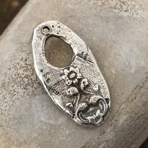 May include: A silver-colored, oval-shaped pendant with a floral design. The pendant has a textured surface and a large oval cutout in the center. The floral design includes a central flower and smaller flowers and leaves.