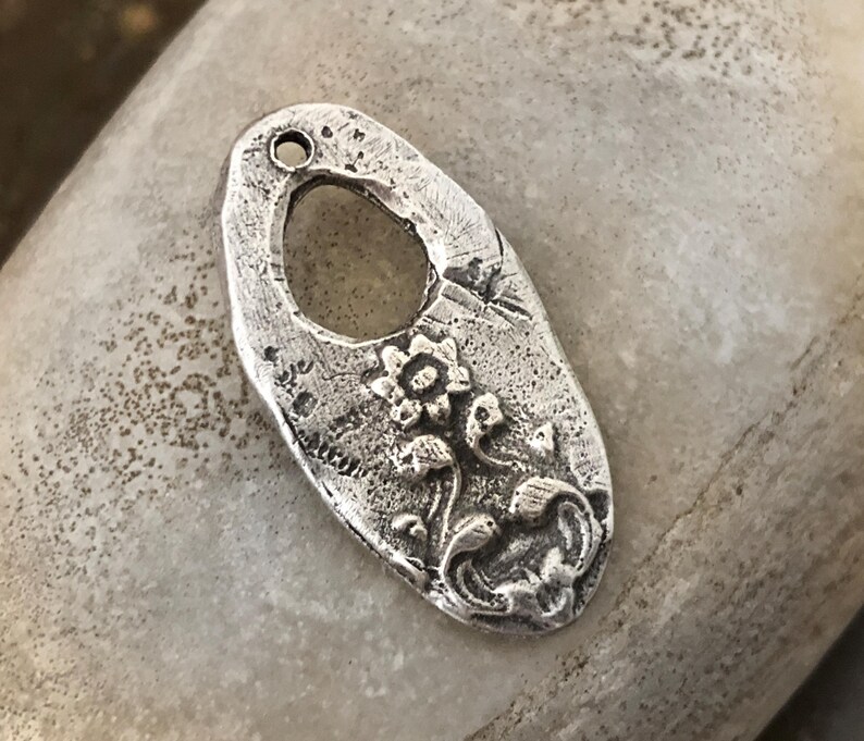 May include: A silver-colored, oval-shaped pendant with a floral design. The pendant has a textured surface and a large oval cutout in the center. The floral design includes a central flower and smaller flowers and leaves.