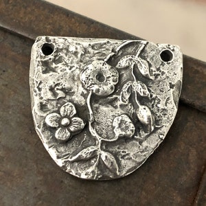 Puede incluir: Un colgante de metal plateado con un dise&ntilde;o floral. El colgante tiene una superficie texturizada y presenta una flor grande, una flor m&aacute;s peque&ntilde;a y hojas. Dos agujeros est&aacute;n en la parte superior para ensartar. El colgante tiene forma de escudo.