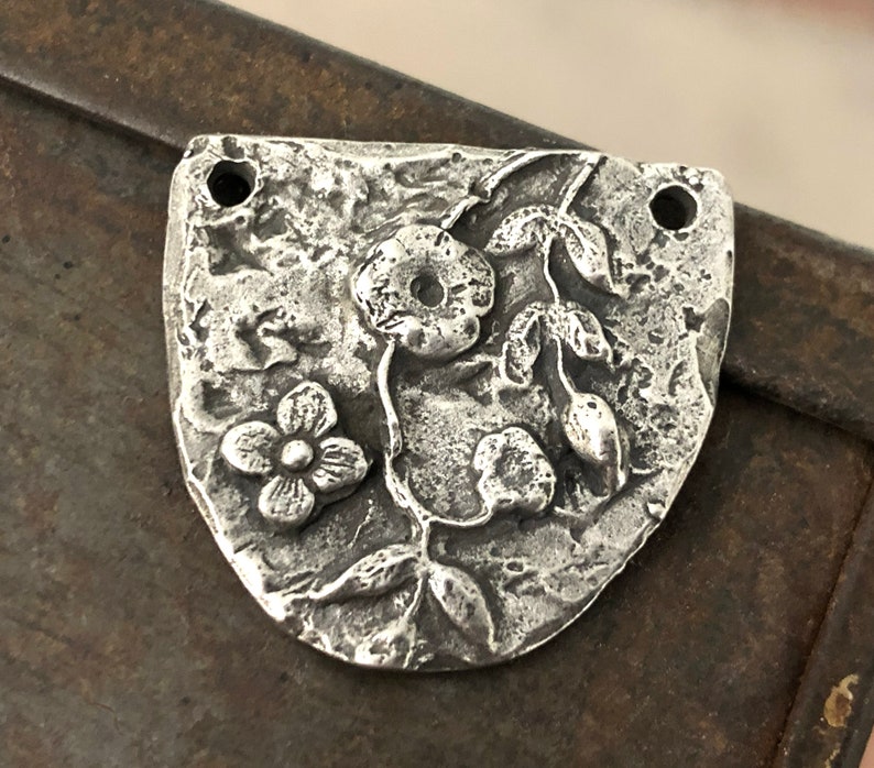Puede incluir: Un colgante de metal plateado con un dise&ntilde;o floral. El colgante tiene una superficie texturizada y presenta una flor grande, una flor m&aacute;s peque&ntilde;a y hojas. Dos agujeros est&aacute;n en la parte superior para ensartar. El colgante tiene forma de escudo.
