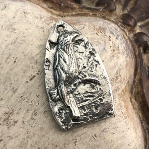 May include: A silver charm pendant featuring a detailed, three-dimensional design of a bird with its wings spread. The charm is shaped like a teardrop and has a textured surface.