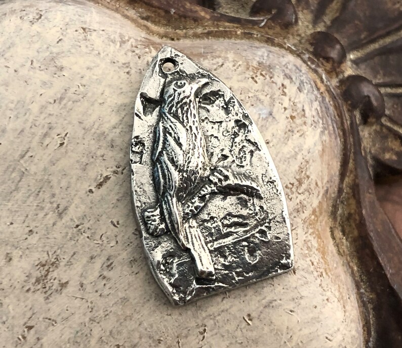 May include: A silver charm pendant featuring a detailed, three-dimensional design of a bird with its wings spread. The charm is shaped like a teardrop and has a textured surface.