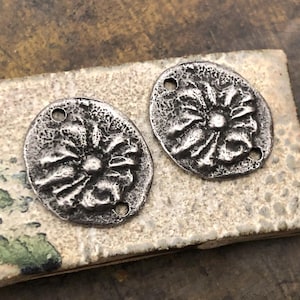 May include: Two antique silver-toned metal flower-shaped pendants. Each pendant has a textured surface with a central flower design and three small holes for stringing. The pendants are oval-shaped and rest on a light-colored, textured surface.