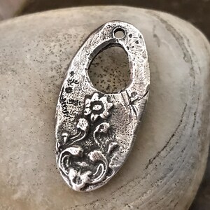 May include: A silver-toned metal pendant with an oval shape and a floral design. The pendant has a textured surface and a hole at the top for hanging. The floral design includes a central flower and flowing stems with leaves.