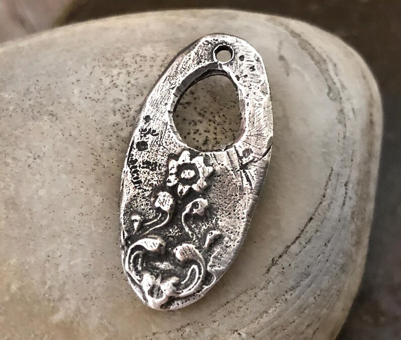 May include: A silver-toned metal pendant with an oval shape and a floral design. The pendant has a textured surface and a hole at the top for hanging. The floral design includes a central flower and flowing stems with leaves.