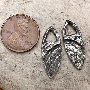 May include: Two silver-toned metal pendants with a textured, wavy design. Each pendant has a small hole at the top for attaching a chain or cord.