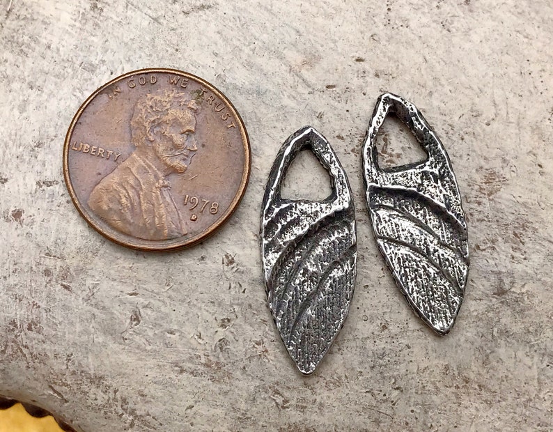 May include: Two silver-toned metal pendants with a textured, wavy design. Each pendant has a small hole at the top for attaching a chain or cord.