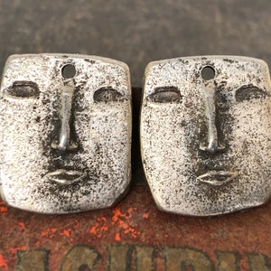 May include: Two silver-toned metal charms shaped like faces with simple features. The charms have a textured surface and a hole at the top for hanging.