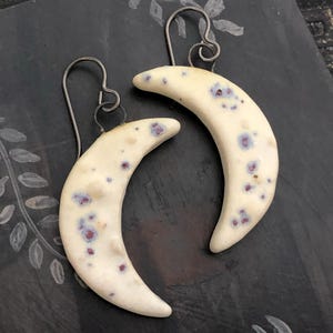 Op de afbeelding: Een paar halve maan oorbellen. De oorbellen zijn gebroken wit met kleine paarse en blauwe spikkels. De oorhaken zijn van donker, gebogen metaal. De oorbellen zijn een accessoire met een hemels thema.