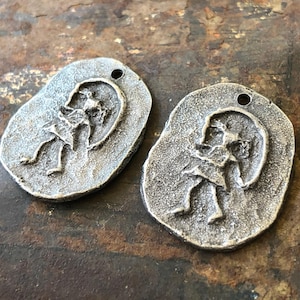 May include: Two silver-toned metal charms shaped like ovals with a raised design of a stylised human figure. The charms have a hole at the top for attaching to a necklace or bracelet.