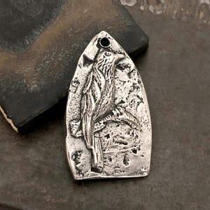 May include: A silver-toned metal pendant with a pointed arch shape, featuring a raised relief design of a bird perched on a branch. The surface has a textured, weathered finish with dark oxidation in the recessed areas and a small hole at the top.