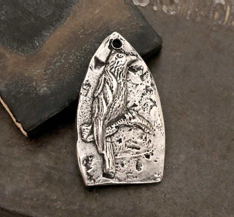 May include: A silver-toned metal pendant with a pointed arch shape, featuring a raised relief design of a bird perched on a branch. The surface has a textured, weathered finish with dark oxidation in the recessed areas and a small hole at the top.
