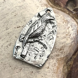 May include: A silver pendant shaped like a bird with a textured surface. The bird is perched on a branch with its wings spread.