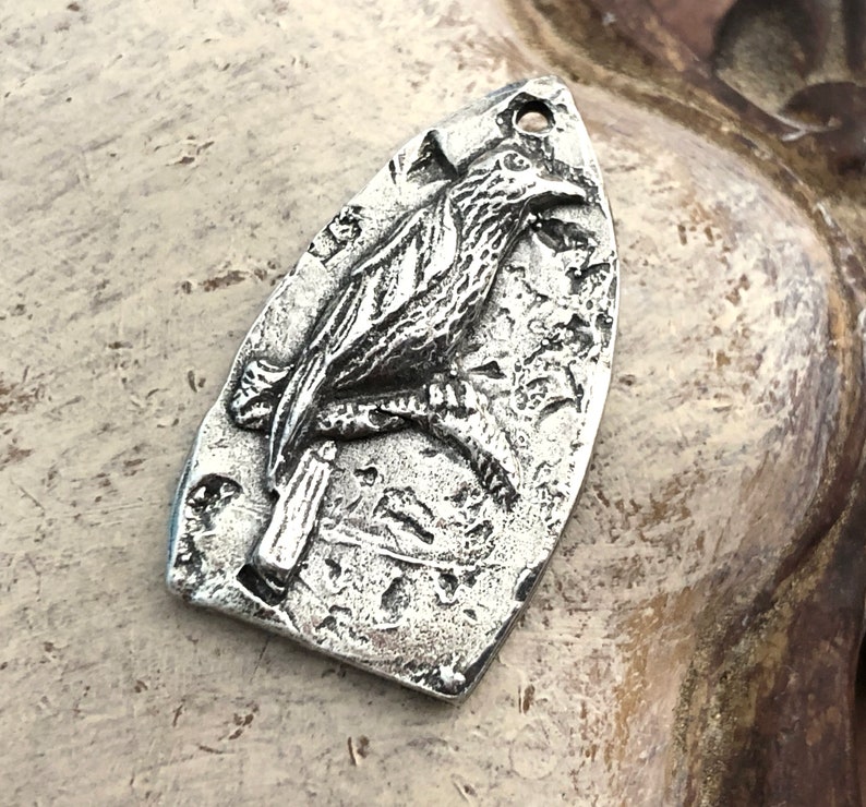 May include: A silver pendant shaped like a bird with a textured surface. The bird is perched on a branch with its wings spread.