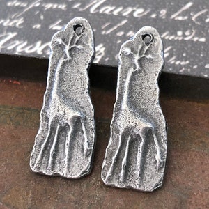 Op de afbeelding: Twee zilverkleurige metalen hangers in de vorm van gestileerde voeten. Elke hanger heeft een getextureerd oppervlak en een gat aan de bovenkant om te rijgen. De hangers worden gepresenteerd op een donker, getextureerd oppervlak.