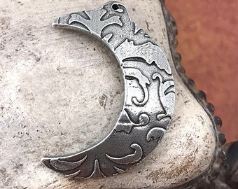 May include: A silver crescent moon pendant with an intricate, textured pattern. The pendant has a hole for threading and is suitable for jewellery making or crafts. The moon shape is a common symbol, often used in pendants.