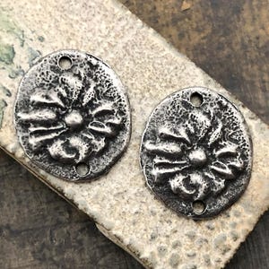 May include: Two antique silver-toned metal pendants. Each pendant is oval-shaped with a textured surface and a floral design in the center. The pendants have two small holes for stringing. The pendants are displayed on a textured, neutral-toned surface.