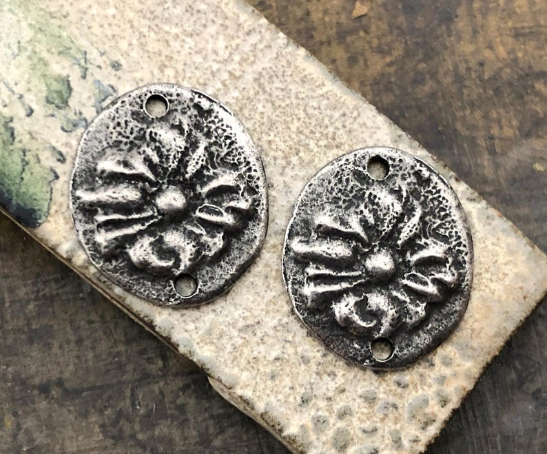 May include: Two antique silver-toned metal pendants. Each pendant is oval-shaped with a textured surface and a floral design in the center. The pendants have two small holes for stringing. The pendants are displayed on a textured, neutral-toned surface.