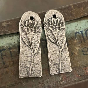 Op de afbeelding: Een paar zilveren oorbellen met een getextureerd, bloemmotief. De oorbellen zijn rechthoekig en hebben een gat aan de bovenkant om te hangen.