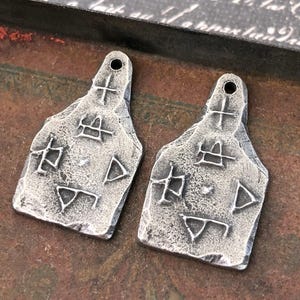 May include: Two silver-toned metal pendants with a textured surface and a hole at the top. Each pendant features an etched design of crosses, squares, and triangles. The pendants are displayed on a dark, rustic background.