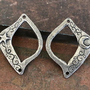 May include: Two silver-toned metal charms in the shape of a diamond with a cutout in the center. One charm features a star and the other features a crescent moon. Both charms have intricate floral designs.