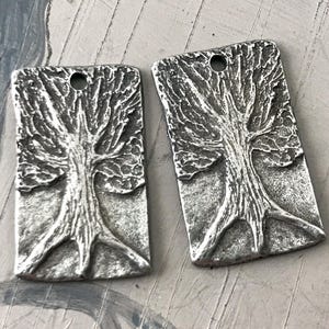 May include: Two silver-toned metal charms shaped like trees with detailed textured branches and trunks. The charms have a hole at the top for attaching to jewelry.