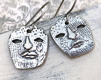 May include: A pair of silver-toned earrings featuring a mask design. Each earring has a square shape with cut-out eye holes, a nose, and a mouth. The masks have textured surfaces and are suspended from silver ear wires.