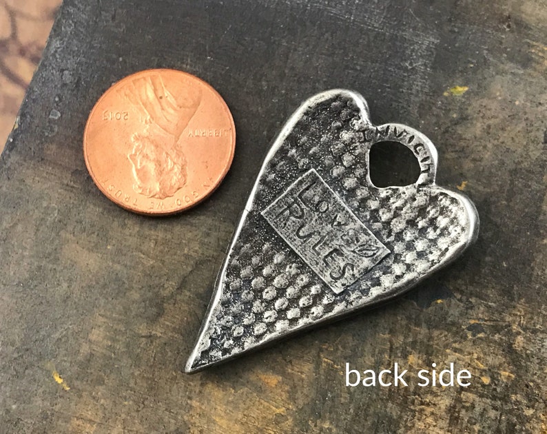 May include: A silver-colored heart-shaped pendant with a textured surface and a rectangular inset that reads "LOVE RULES". A U.S. penny is next to the pendant for size comparison. The pendant has a small hole at the top.