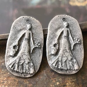 Op de afbeelding: Twee zilverkleurige metalen bedels in de vorm van een ovaal met een reliëfdesign van een vrouw die een emmer en een tak draagt. De bedels hebben een gat aan de bovenkant om te hangen.