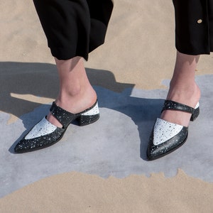 May include: A pair of black and white heeled mules with a pointed toe. The shoes have a white base with black speckles and a black strap across the top.