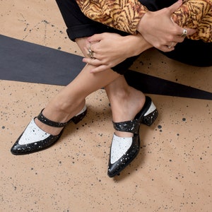 May include: A pair of black and white heeled mules with a splatter paint design. The shoes have a pointed toe and a strap across the top of the foot.