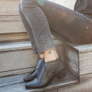 May include: Black leather ankle boots with a low heel. The boots are paired with dark grey denim jeans. A black top and a white fluffy jacket complete the outfit. The person is sitting on wooden steps.