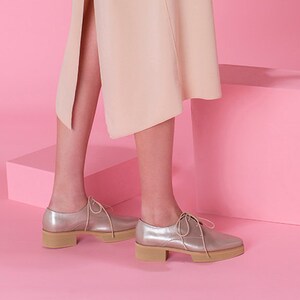 May include: A person wearing a light pink sleeveless dress and silver shoes holds a potted plant with long, green leaves against a pink background.