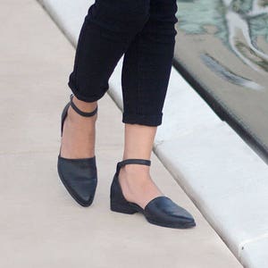 May include: A pair of black ankle strap flats with a pointed toe. The shoes are worn by a person wearing black jeans.