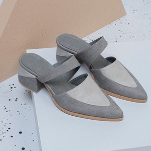 May include: A pair of gray suede heeled mules with a pointed toe and a strap across the top of the foot. The shoes are on a white platform.