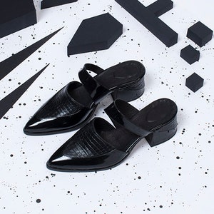 Puede incluir: Dos mules negras de tac&oacute;n de cuero con punta puntiaguda y una tira en la parte superior. Los zapatos est&aacute;n sobre una superficie blanca con formas geom&eacute;tricas negras y salpicaduras de pintura negra.