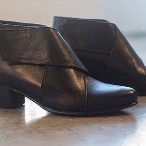 May include: A pair of black leather ankle boots. The boots have a distinctive design with a folded-over flap and a low, stacked heel. The boots are displayed on a light-coloured surface.