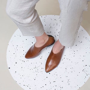 May include: A pair of brown leather slip-on shoes with pointed toes. The shoes are on a white surface with black dots.