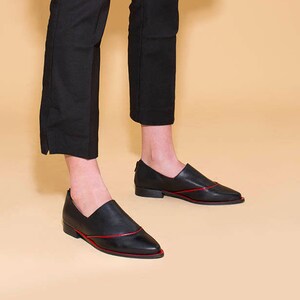 Puede incluir: Zapatos de vestir negros de cuero con un detalle de costura roja. Los zapatos tienen una punta puntiaguda y un tac&oacute;n bajo.