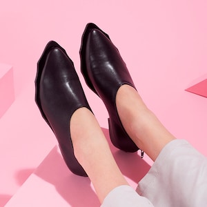 May include: A pair of black leather pointed-toe shoes with a unique scalloped edge design. The shoes are worn by a person with white pants on a pink background.